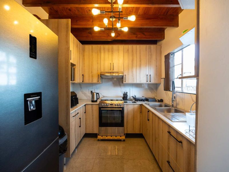 Modern kitchen featuring light wood cabinetry, exposed timber ceiling beams, stainless steel appliances, and a double sink with a tiled backsplash.