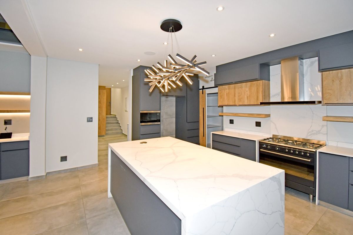 Modern kitchen featuring a large quartz-topped island, dark grey and wood-tone cabinetry, high-end gas range, and integrated lighting, leading to a separate scullery area.