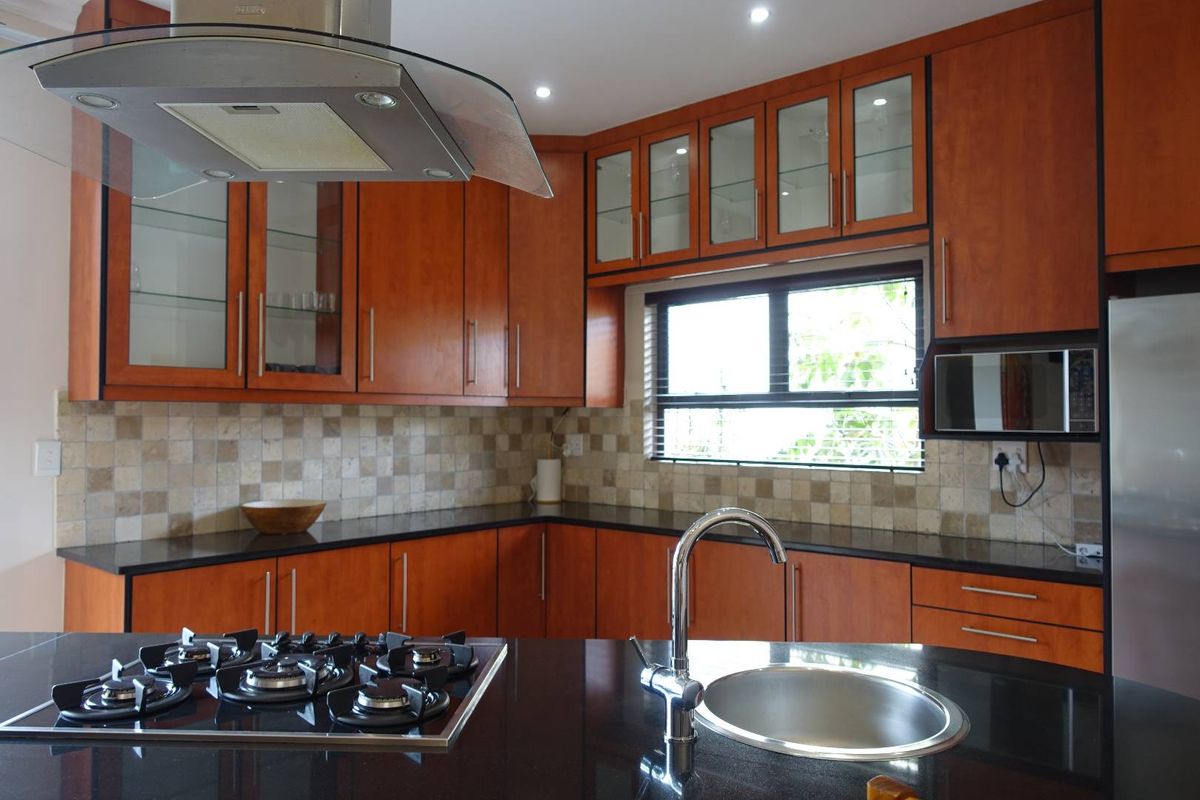 Modern kitchen featuring warm-toned wooden cabinetry, dark granite countertops, a central island with a gas hob and sink, and a glass extractor fan.