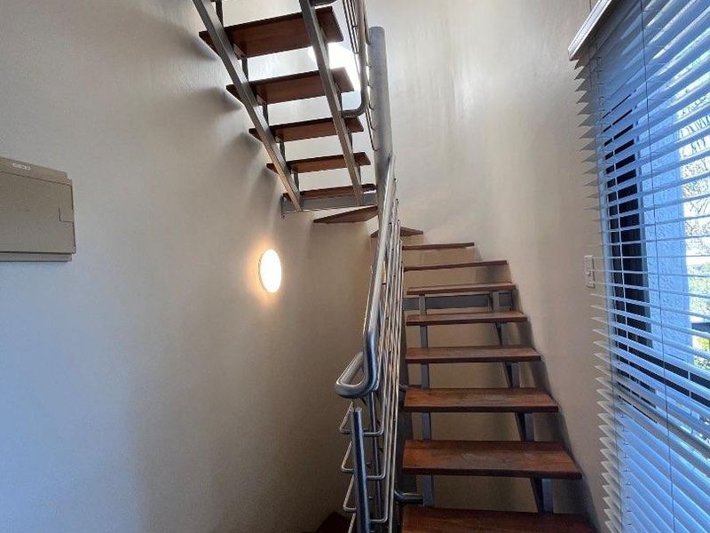 A modern staircase featuring timber treads and a stainless steel balustrade, set against a neutral wall with slate flooring and venetian blinds.