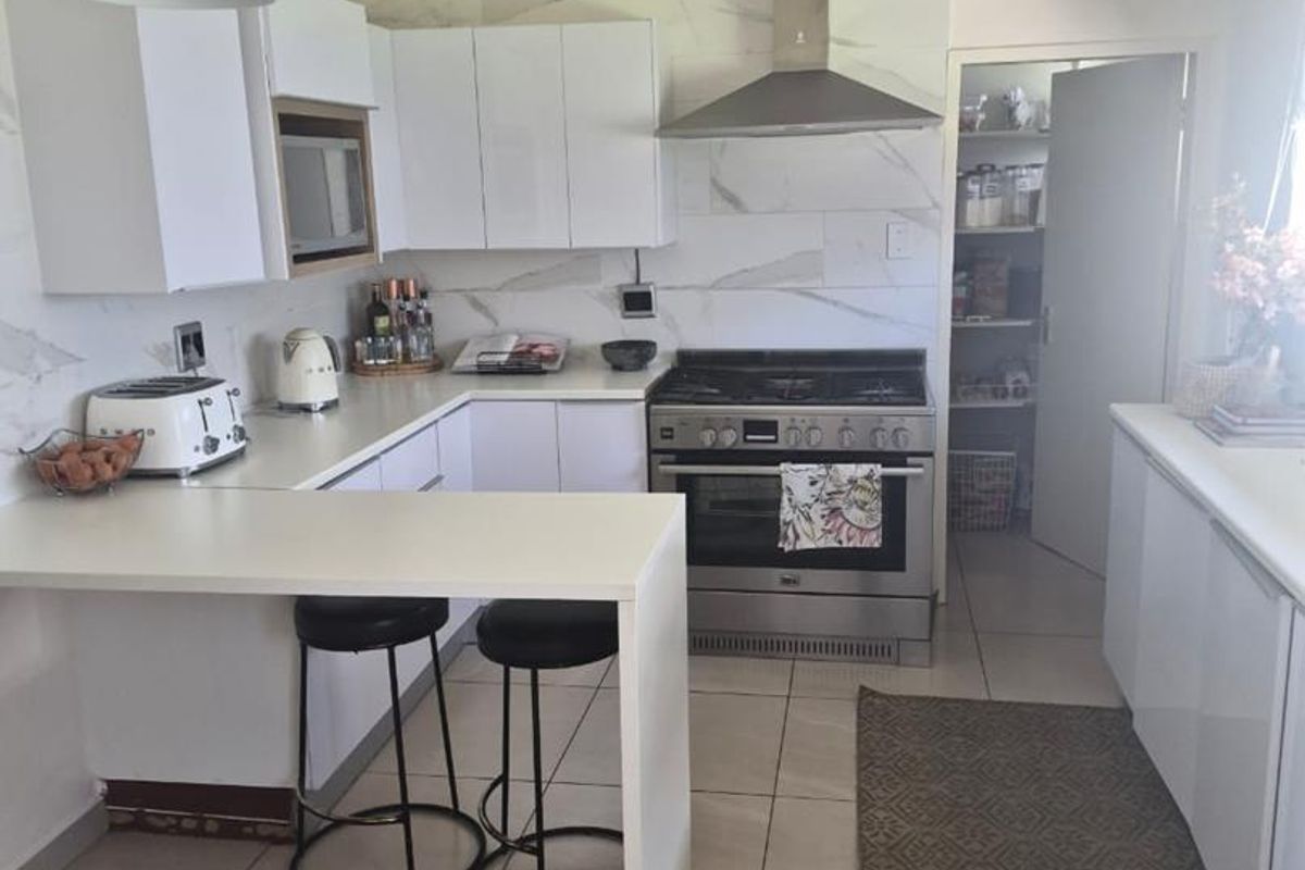 Modern kitchen featuring white cabinetry, marble-look splashback, stainless steel gas stove with extractor fan, and an integrated breakfast bar, with an adjacent walk-in pantry.