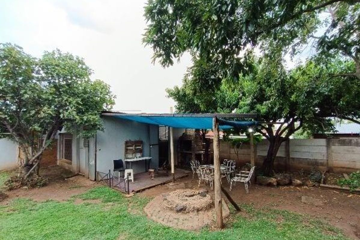 Rear garden area featuring a covered patio, built-in braai, and a central fire pit, surrounded by mature trees and boundary walling.