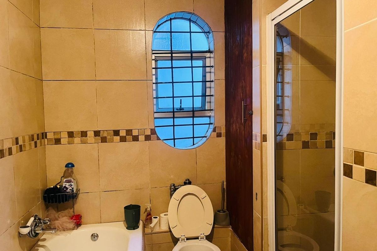 A compact bathroom featuring a bathtub, separate shower enclosure, and toilet, finished with dated wall tiling and a unique arched window with security bars.