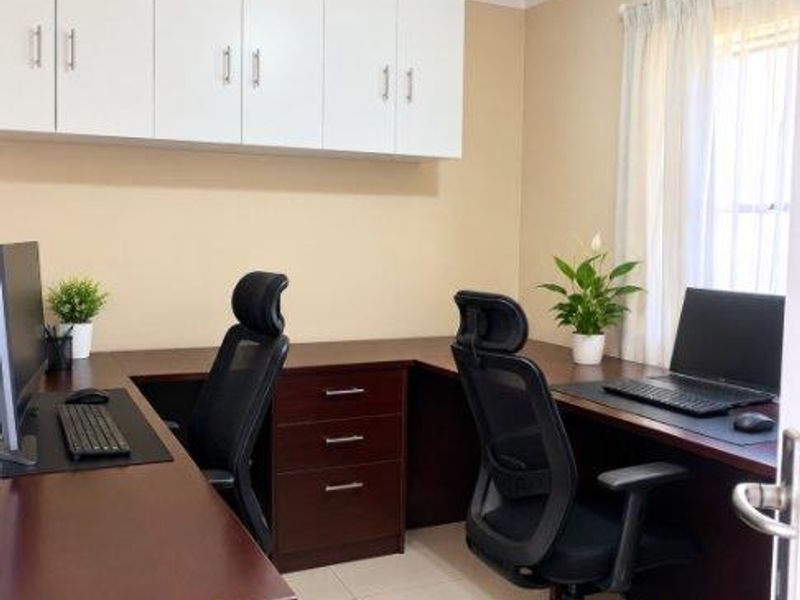 A functional home office featuring built-in dark wood desk units, ample white overhead storage cabinetry, and neutral tiled flooring.