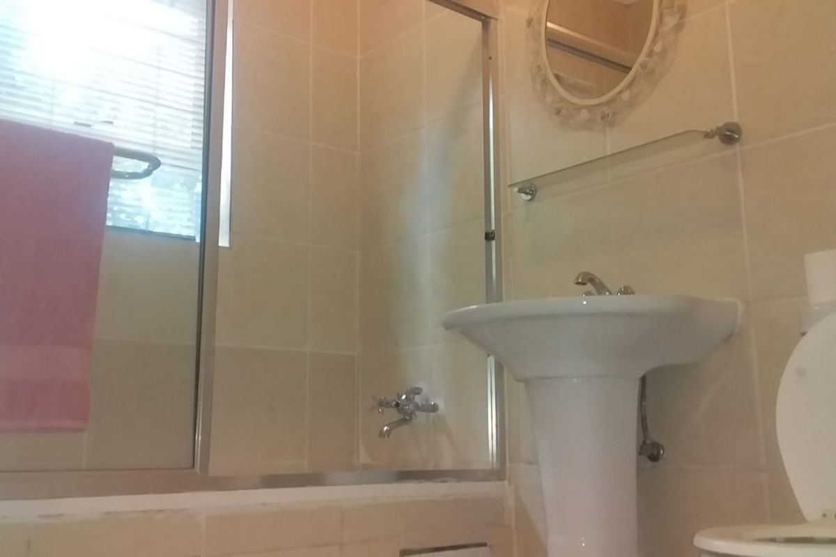 A dated bathroom featuring a pedestal sink, a shower-over-bath combination with a glass screen, and neutral wall tiling.