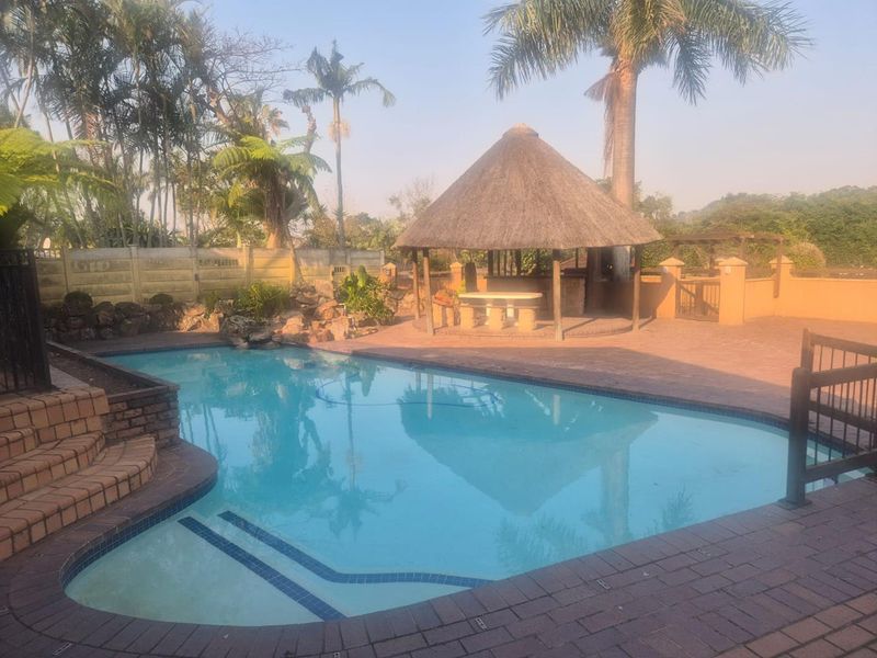 Outdoor pool area featuring a swimming pool, brick paved surround, and a traditional thatch lapa, set within a landscaped garden environment.