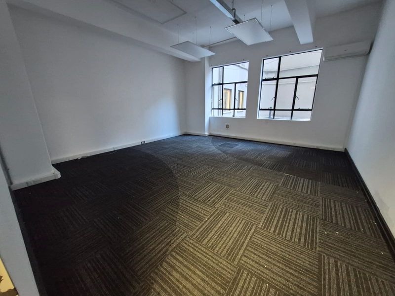 An empty office space featuring carpet tile flooring, large multi-pane windows for natural light, and a wall-mounted air conditioning unit.