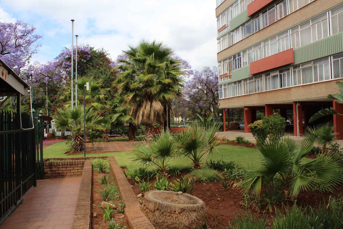 A view of the apartment complex exterior featuring a landscaped garden, paved walkways, and established trees adjacent to the building facade.
