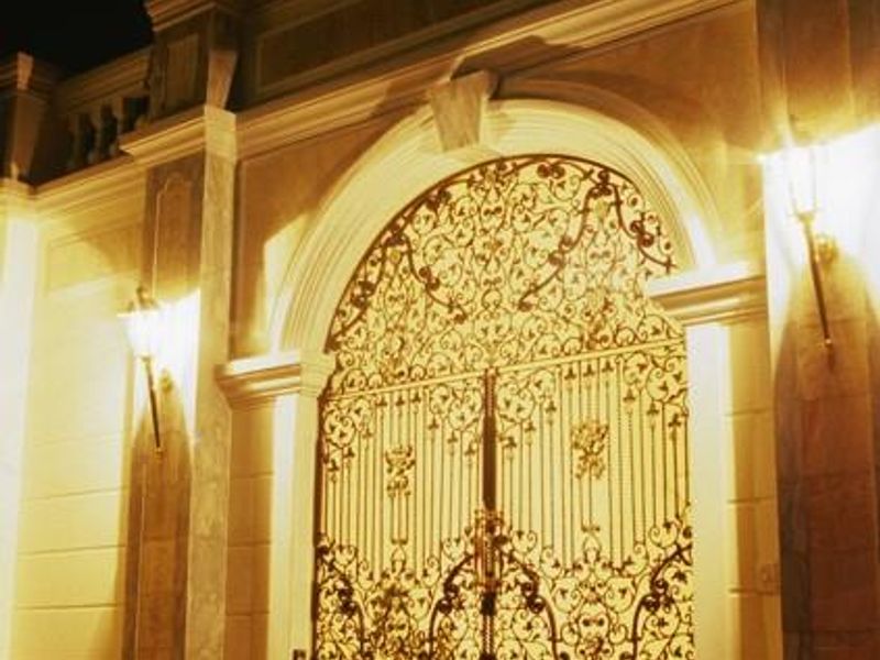 A grand, illuminated front entrance featuring an ornate wrought iron double gate set within a classic stone archway.
