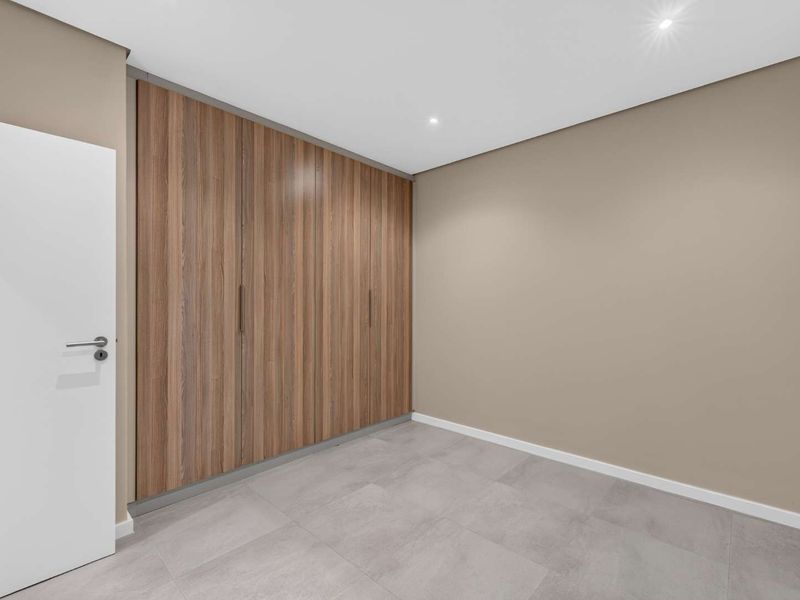 A clean, modern bedroom featuring neutral wall tones, large-format floor tiles, and floor-to-ceiling built-in wooden cupboards with recessed lighting.