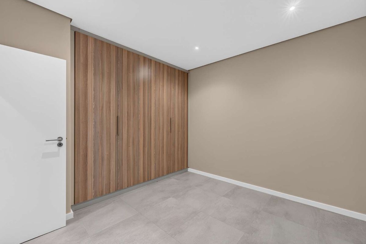 A clean, modern bedroom featuring neutral wall tones, large-format floor tiles, and floor-to-ceiling built-in wooden cupboards with recessed lighting.