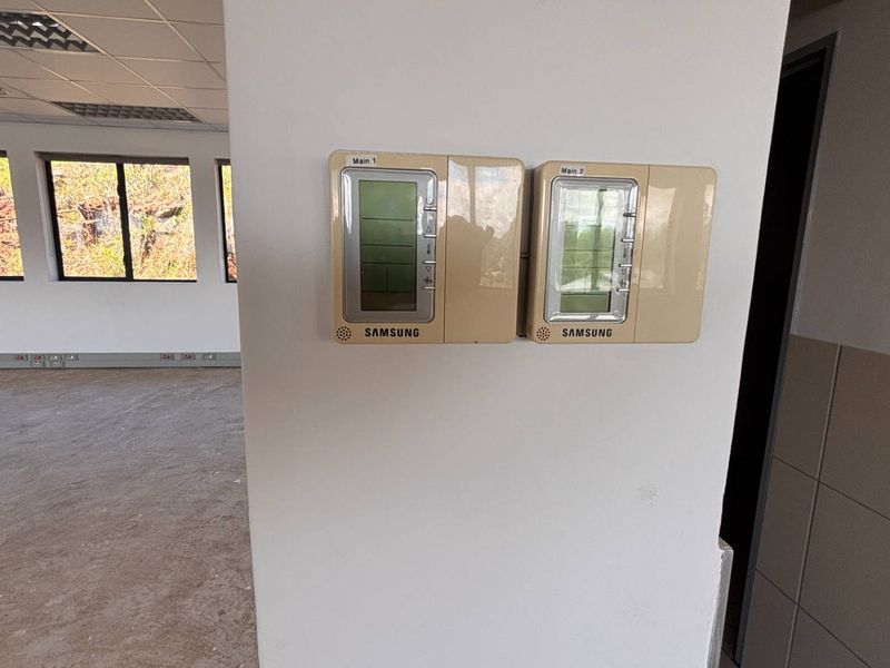 An open-plan office space featuring suspended ceilings, recessed lighting, aluminum-framed windows, and wall-mounted HVAC control panels.