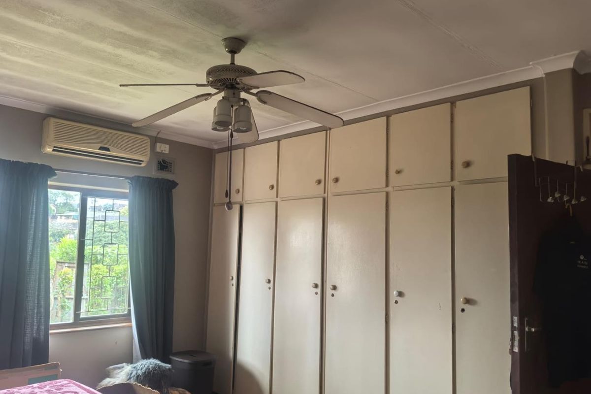 Bedroom featuring extensive built-in cupboards, a ceiling fan, and a wall-mounted air conditioning unit with security bars on the window.