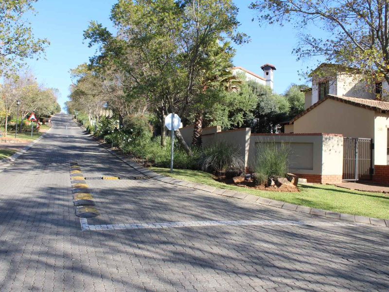 A view of the paved residential street approach, featuring speed bumps, mature trees, and the boundary wall of the property.