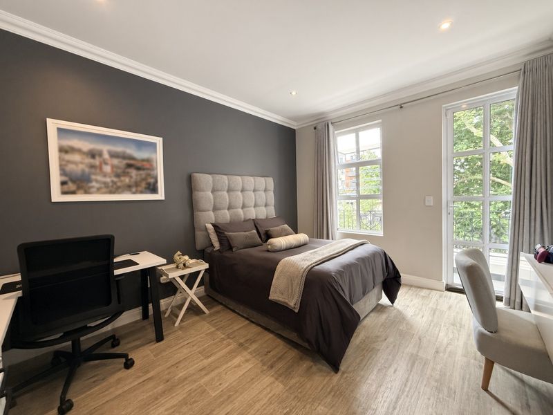 A well-maintained bedroom featuring modern laminate flooring, crown molding, recessed lighting, and large windows providing natural light and balcony access.