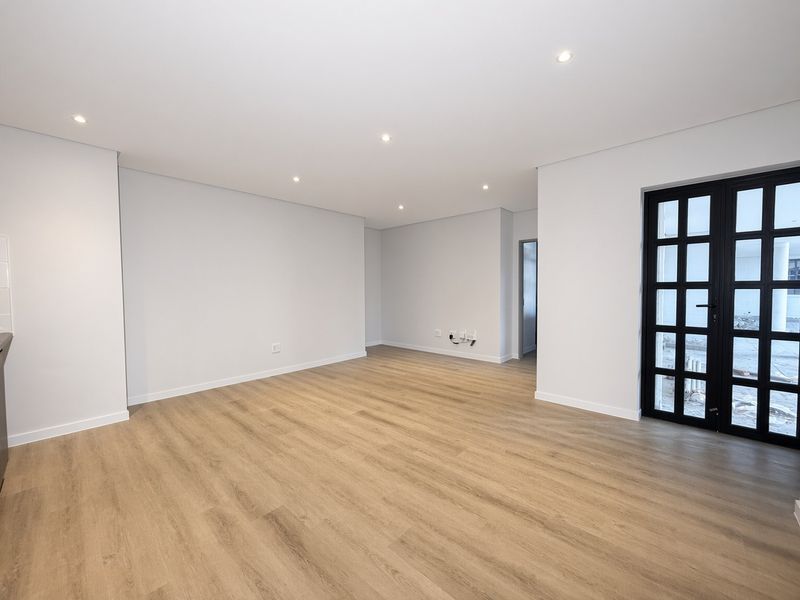 A bright, modern, and empty living area featuring light wood-look laminate flooring, recessed ceiling lighting, and a stylish black-framed glass door leading outdoors.