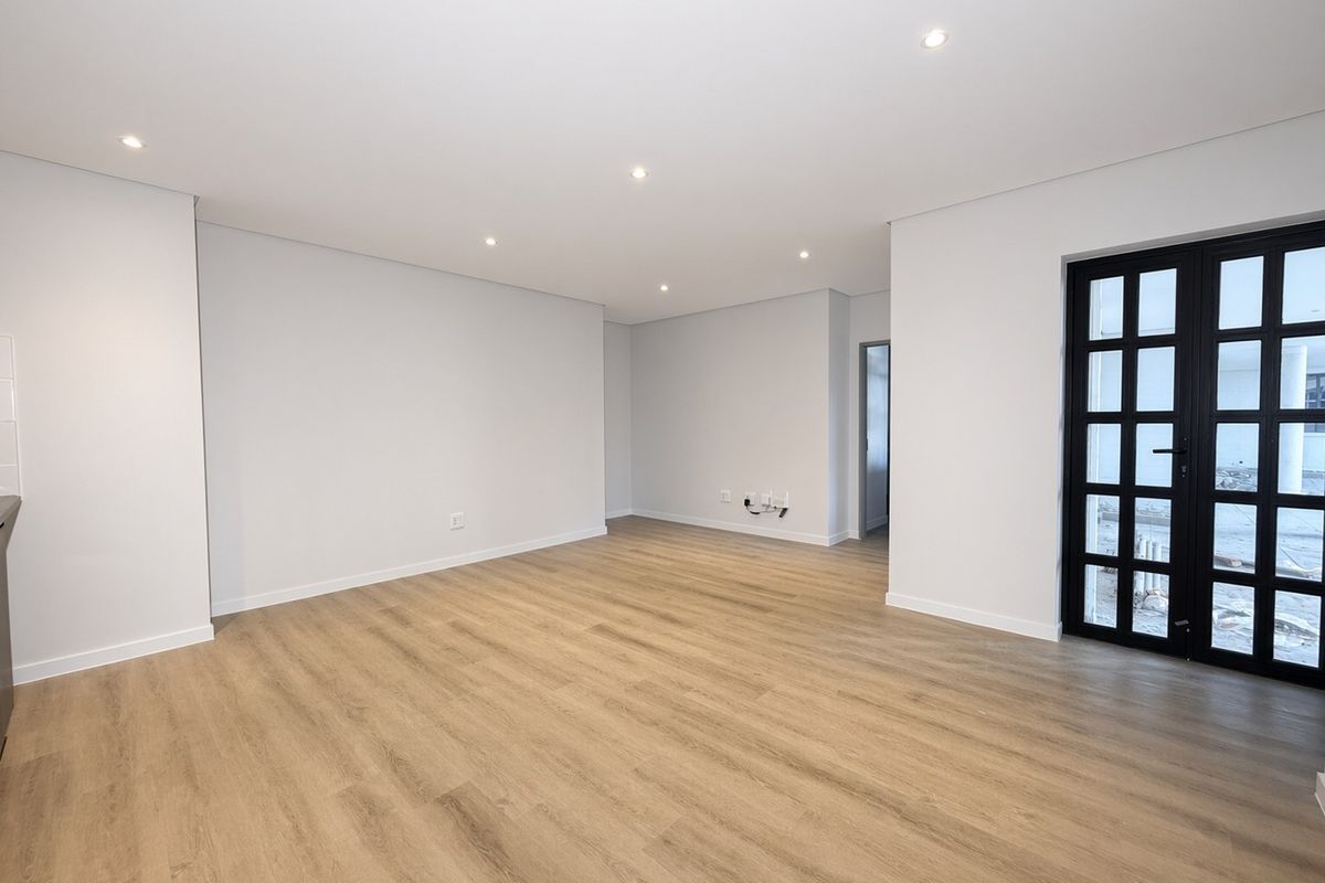 A bright, modern, and empty living area featuring light wood-look laminate flooring, recessed ceiling lighting, and a stylish black-framed glass door leading outdoors.
