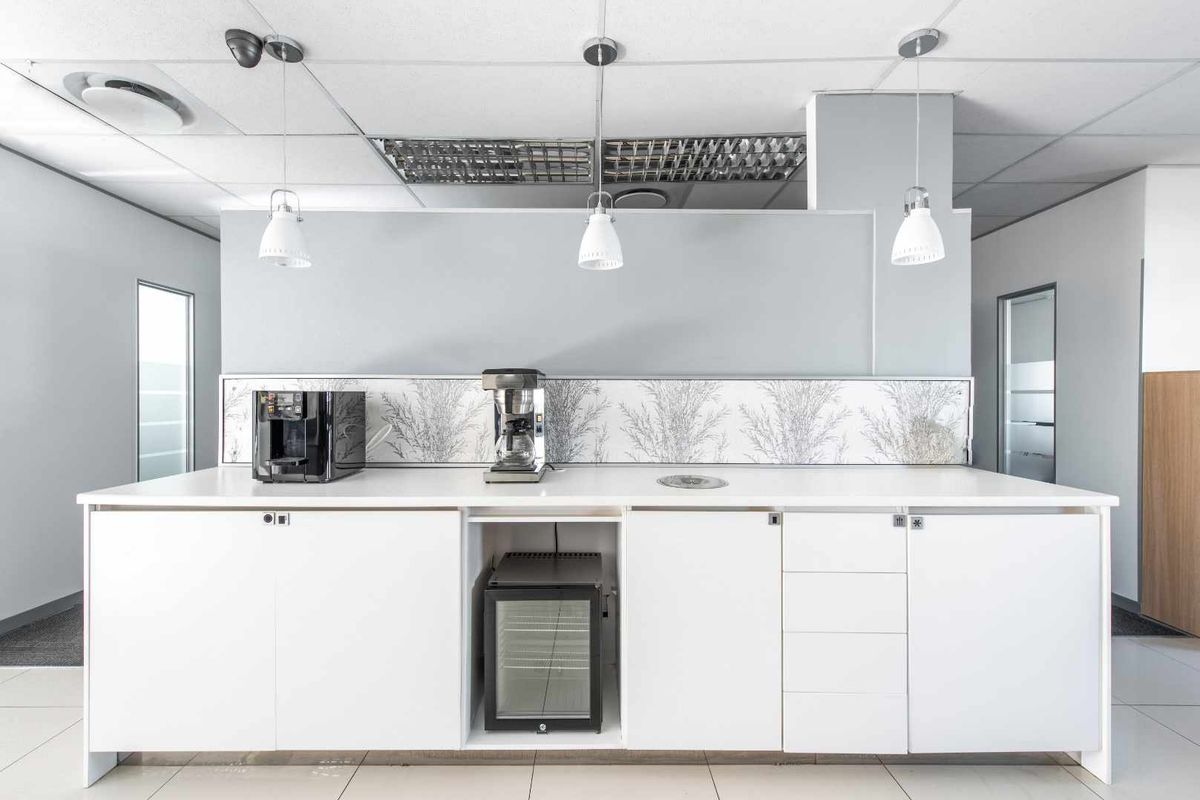 A modern office kitchenette featuring white cabinetry, a sleek countertop, integrated under-counter beverage cooler, and stylish pendant lighting.