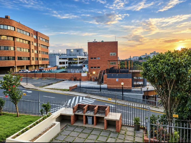Outdoor entertainment area featuring a built-in brick braai, paved patio, and landscaped garden with views of the surrounding apartment complex and sunset.