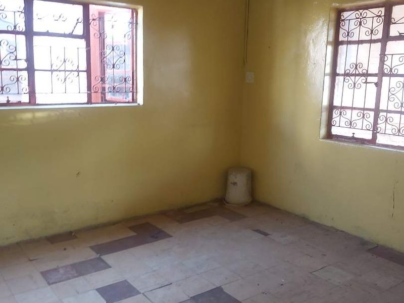An empty bedroom featuring tiled flooring, yellow painted walls, and steel security bars on the windows, requiring cosmetic updates and floor repairs.