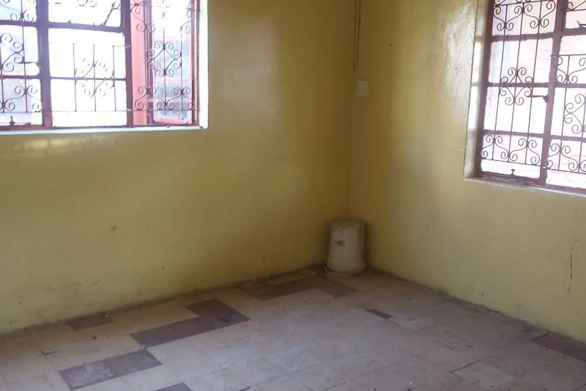 An empty bedroom featuring tiled flooring, yellow painted walls, and steel security bars on the windows, requiring cosmetic updates and floor repairs.