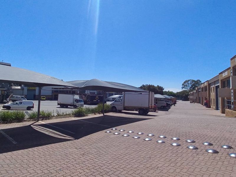 Industrial complex exterior featuring a paved driveway, shade cloth carports, and multiple roller shutter doors for warehouse access.