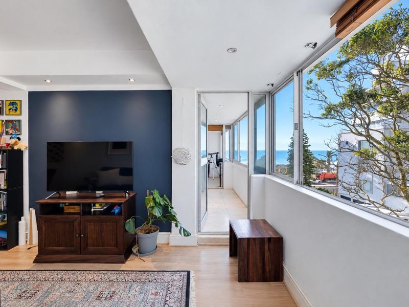 Bright living room featuring timber flooring, an accent wall, and large windows providing natural light and access to an enclosed balcony with ocean views.