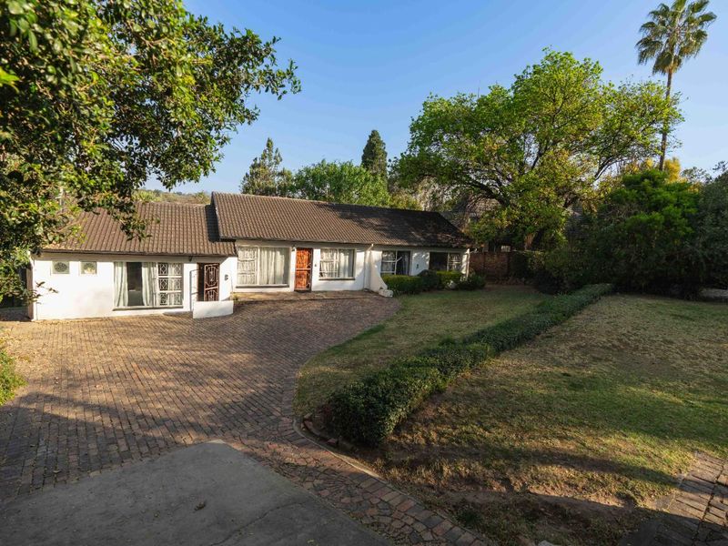 Front view of a single-story house featuring a brick-paved driveway, established garden with mature trees, and a tiled roof.