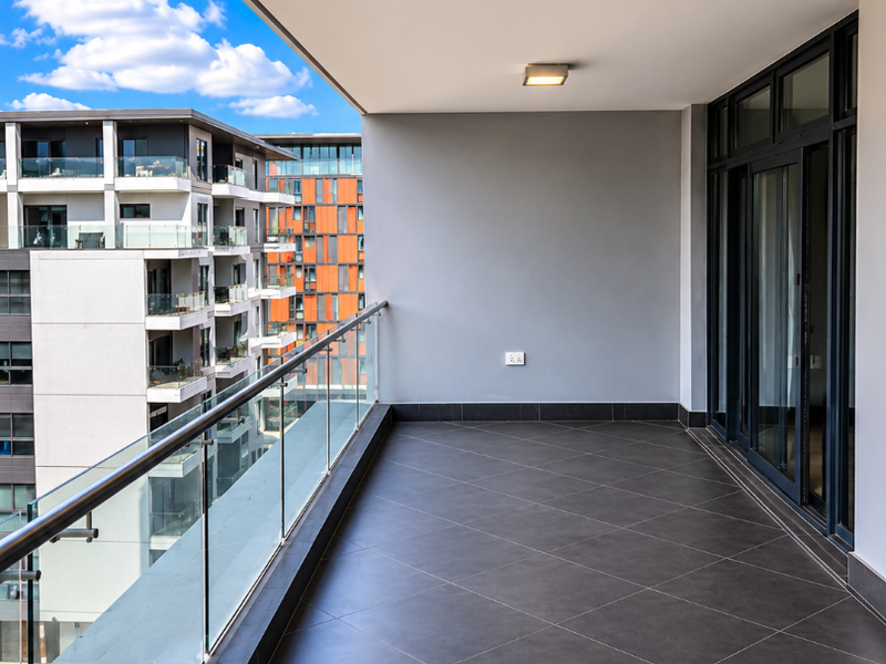 A modern, covered balcony featuring dark tiled flooring, a sleek glass balustrade, and sliding glass door access to the apartment interior.
