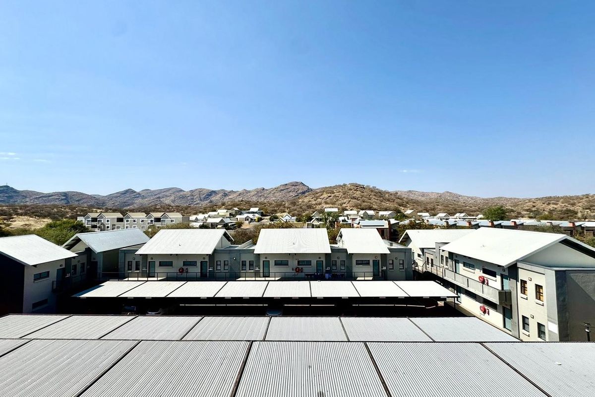 An elevated aerial view of a residential apartment complex featuring corrugated metal roofs and covered parking structures set against a mountain landscape.