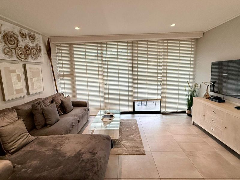 A well-maintained living room featuring large format floor tiles, recessed lighting, and a sliding glass door with venetian blinds leading to an outdoor area.