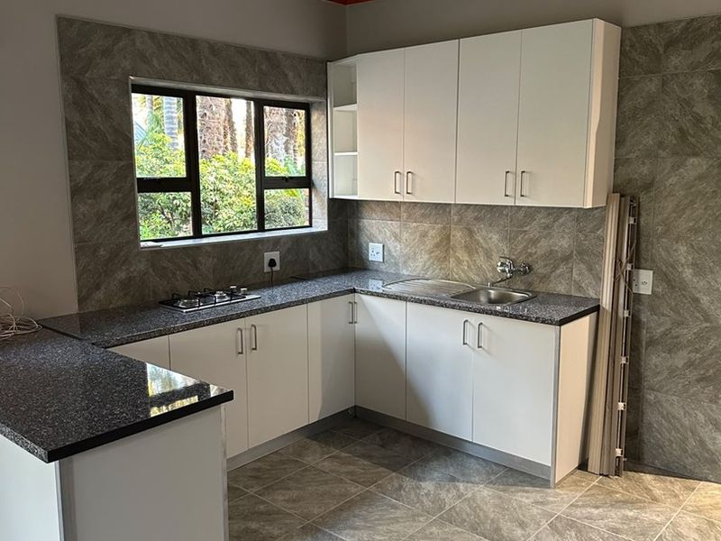 A clean, modern kitchen featuring granite countertops, built-in hob, stainless steel sink, and exposed timber ceiling beams with neutral tiled finishes.