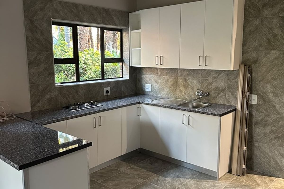 A clean, modern kitchen featuring granite countertops, built-in hob, stainless steel sink, and exposed timber ceiling beams with neutral tiled finishes.