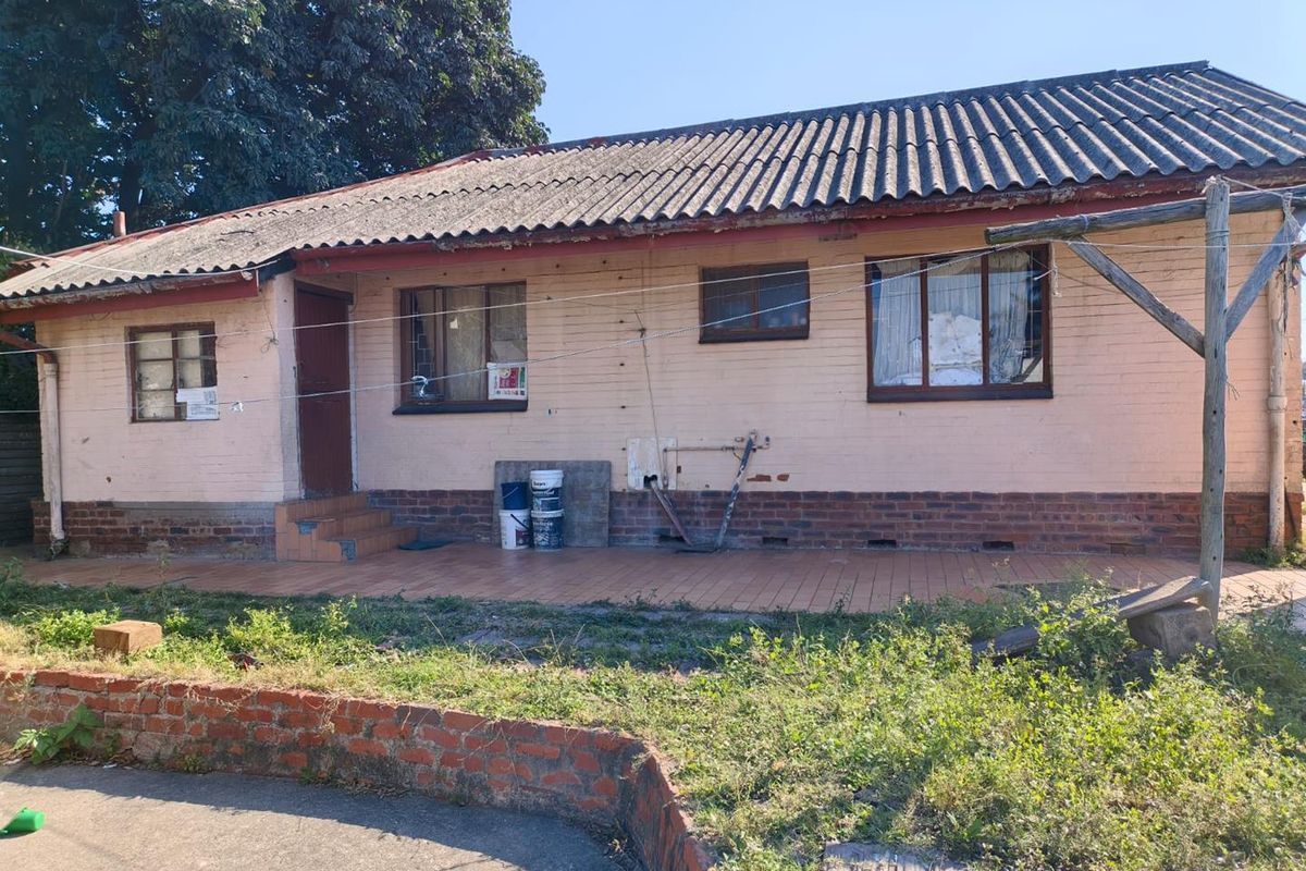 Rear view of a single-story brick house featuring a tiled patio, corrugated roof, and surrounding garden area in need of maintenance.