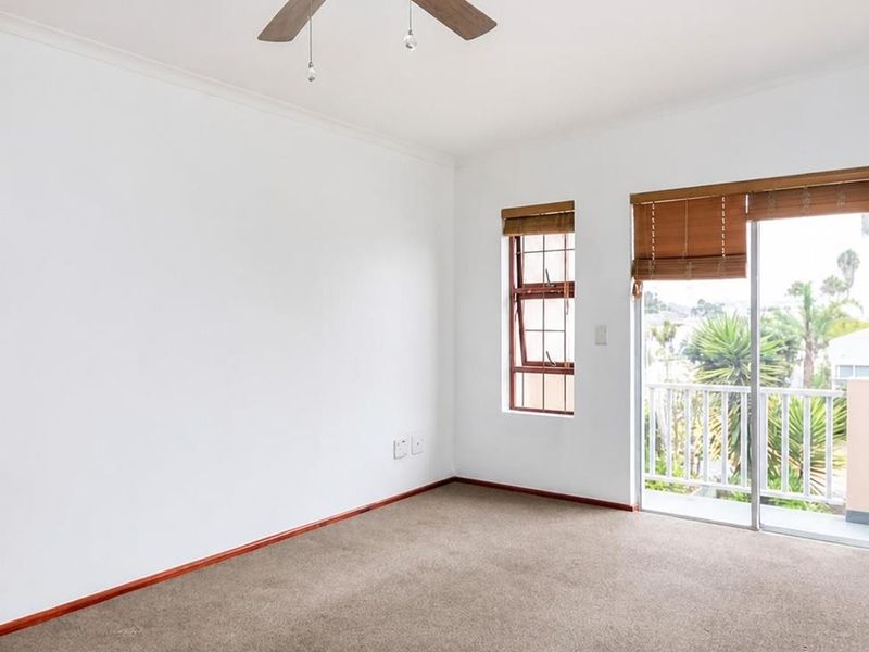 A bright, empty bedroom featuring neutral carpeting, a ceiling fan, and a sliding glass door providing access to a balcony with outdoor views.