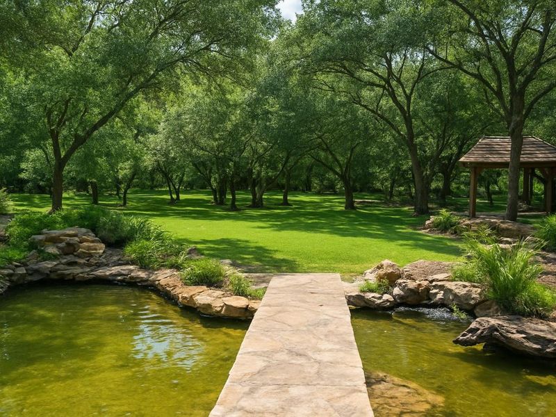 A serene garden landscape featuring a stone footbridge over a rock-lined pond, leading to a lush green lawn and a charming wooden gazebo.