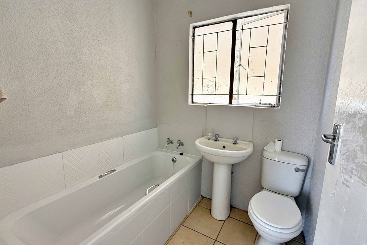 A functional bathroom featuring a standard bathtub, pedestal basin, and toilet, with a window providing natural light and security bars.