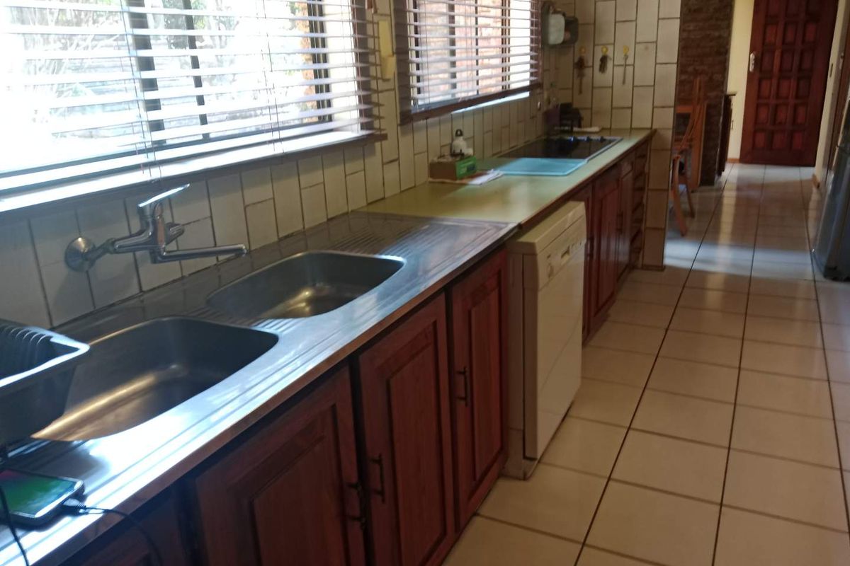 A functional kitchen featuring wooden cabinetry, a double stainless steel sink, tiled splashbacks, and tiled flooring with a view through horizontal blinds.