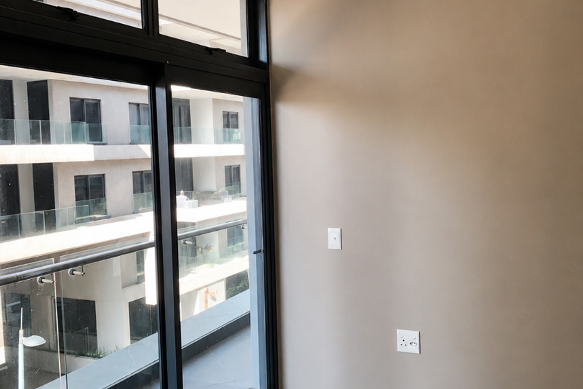 A bright, empty living area featuring large sliding glass doors that open onto a balcony, with neutral walls and tiled flooring.
