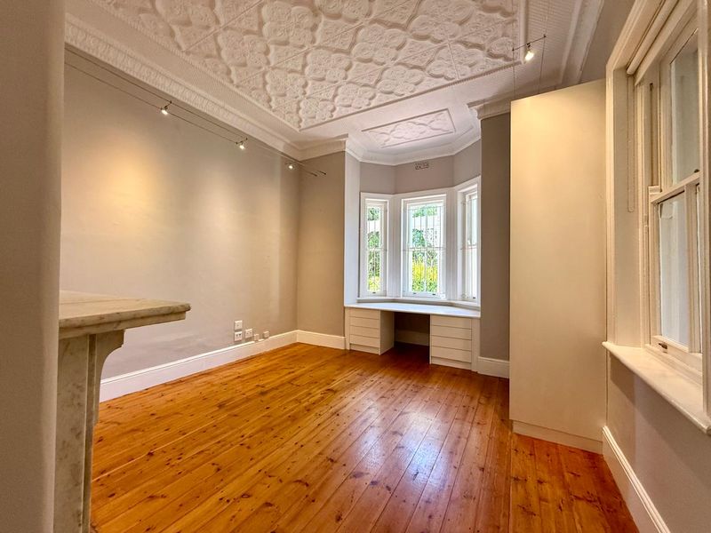 Spacious bedroom featuring original timber flooring, an ornate pressed metal ceiling, a marble fireplace mantel, and a built-in desk within a charming bay window.