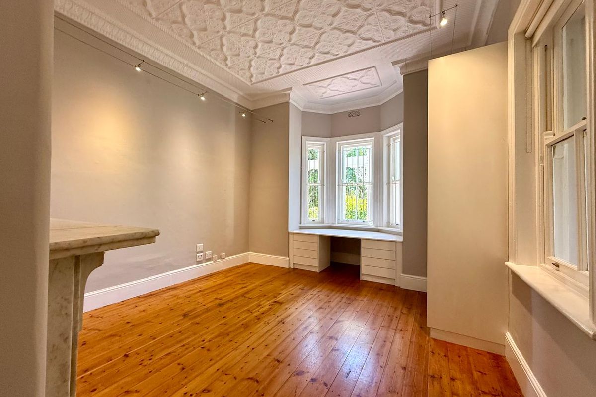 Spacious bedroom featuring original timber flooring, an ornate pressed metal ceiling, a marble fireplace mantel, and a built-in desk within a charming bay window.