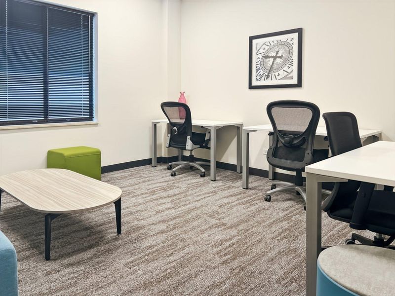 A well-maintained office space featuring neutral carpet flooring, clean white walls, and large windows with fitted venetian blinds.