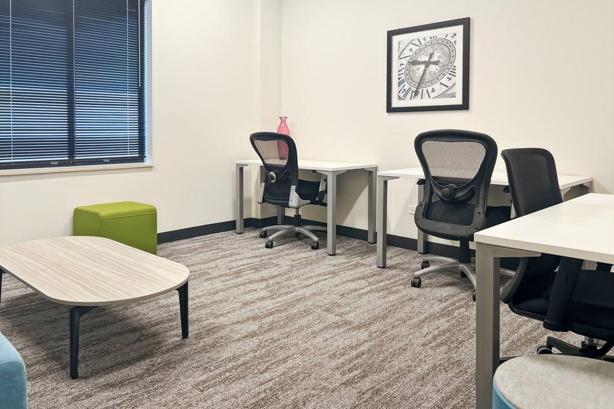 A well-maintained office space featuring neutral carpet flooring, clean white walls, and large windows with fitted venetian blinds.