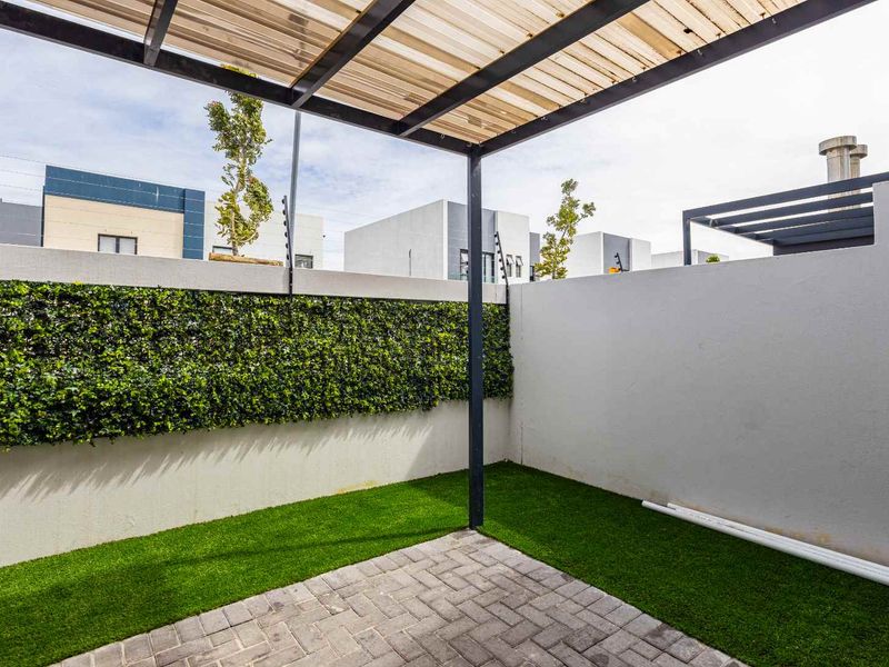 A neat, low-maintenance outdoor patio area featuring paved flooring, artificial turf, and a covered pergola structure, enclosed by a secure boundary wall.