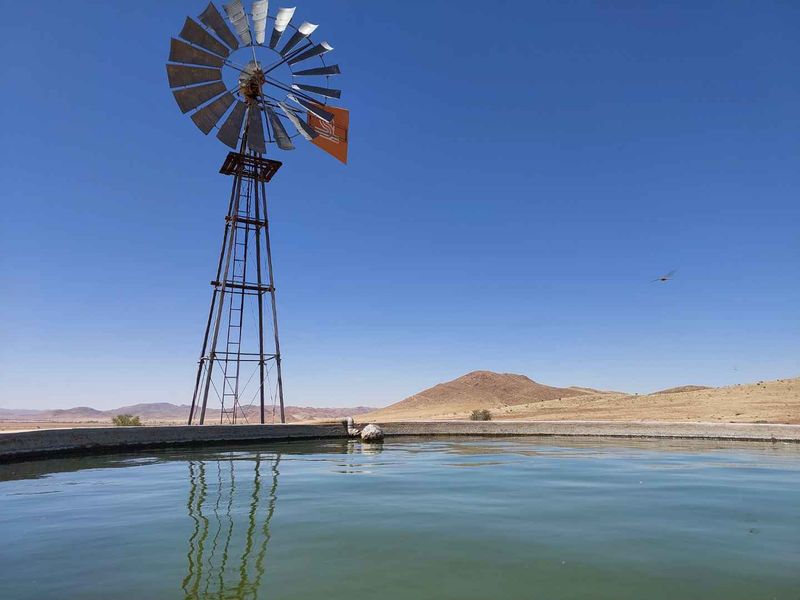 A traditional farm windmill standing beside a concrete water reservoir, set against a vast arid landscape with distant hills.