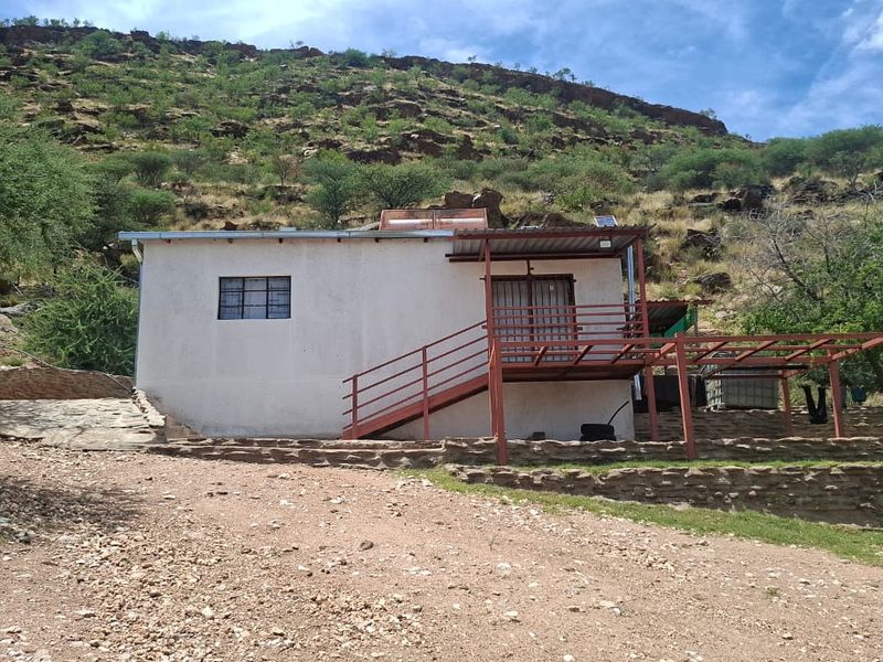 Rear view of a small holding dwelling featuring a raised deck, metal staircase, and stone retaining wall set against a natural hillside.