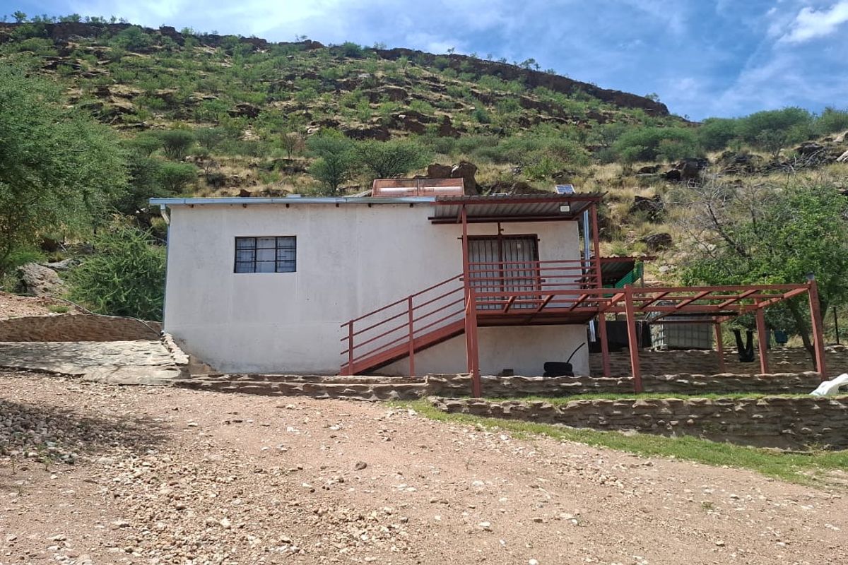 Rear view of a small holding dwelling featuring a raised deck, metal staircase, and stone retaining wall set against a natural hillside.