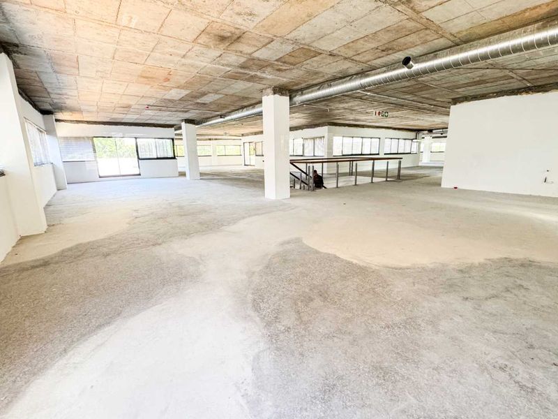 Expansive open-plan commercial space featuring an industrial aesthetic with exposed concrete ceilings, ducting, and large windows providing ample natural light.