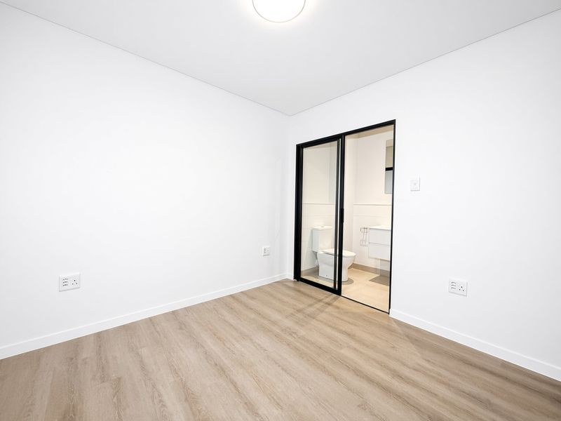 A bright, modern bedroom featuring light wood-look laminate flooring, crisp white walls, and a sleek black-framed sliding door leading to an en-suite bathroom.