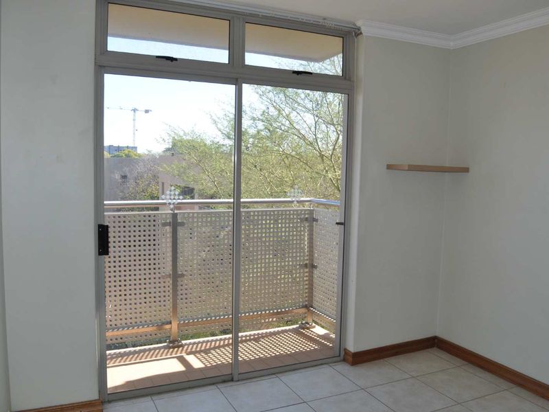 A bright, empty bedroom featuring tiled floors, a floating shelf, and a sliding glass door leading to a private balcony with a metal balustrade.